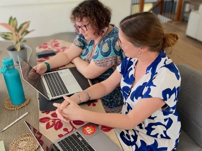 Celia and Michelle sitting at a desk working on a retreat project together using their laptops.